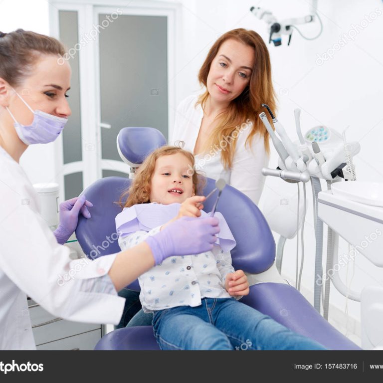 Female dentist in uniform with assistant showing a tool to little girl sitting in chair in a hospital. Dentist and child in cabinet. Dentist office.